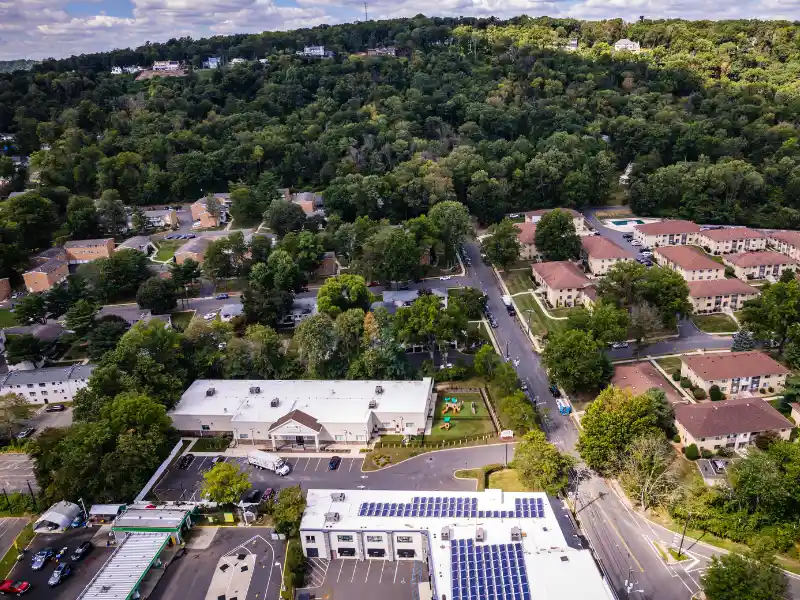 Aerial view of a suburban neighborhood with buildings, roads, and a forested hill in the background.