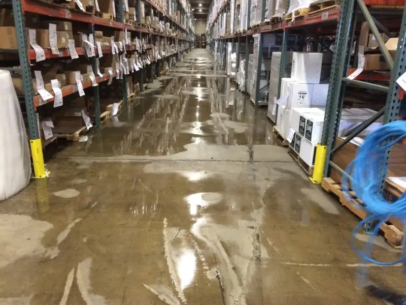 A warehouse aisle with wet, reflective concrete floor and shelves stocked with boxes on both sides.