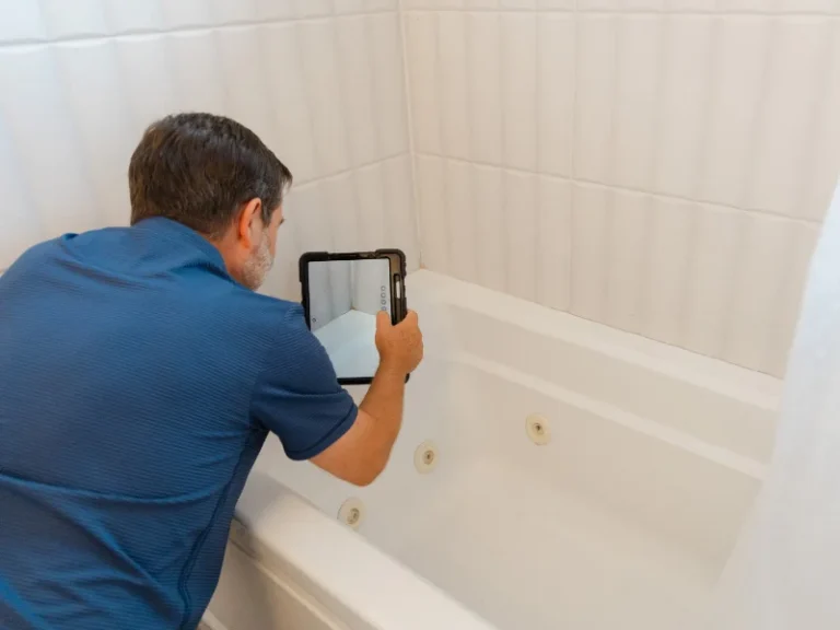 Man in blue shirt inspecting a bathtub with a tablet in a tiled bathroom.