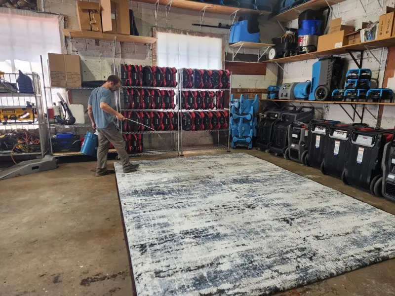 Man spraying a large rug in a storage room filled with equipment and shelves.