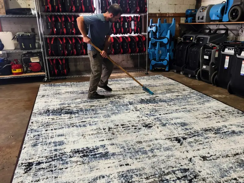 Man cleaning a large patterned rug with a mop in a storage room.