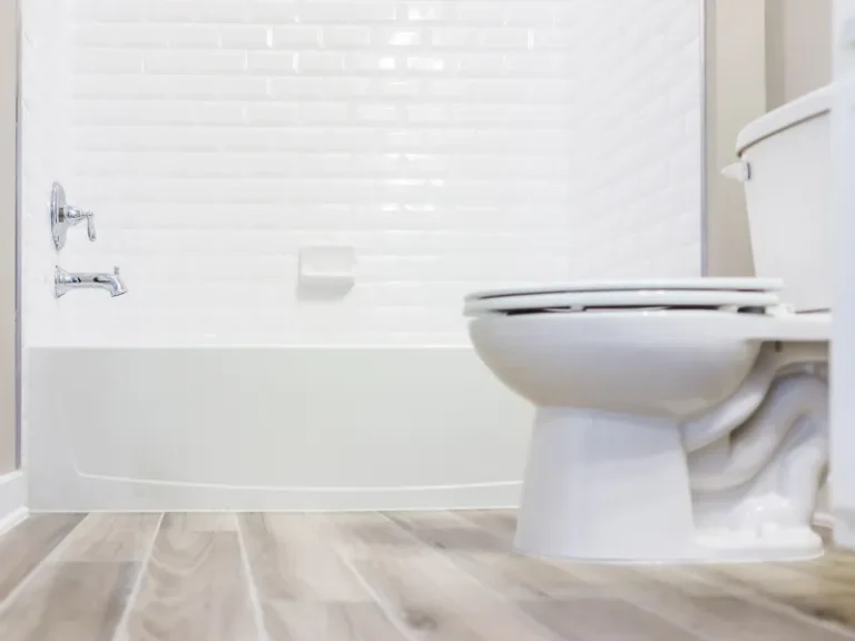 White toilet with closed lid next to a white bathtub with tiled walls and wood-look floor.